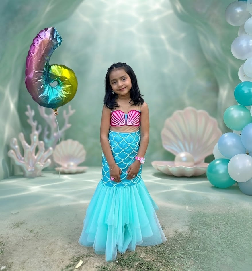 Young girl in a mermaid costume standing in an underwater-themed setting with balloons and shell decorations.